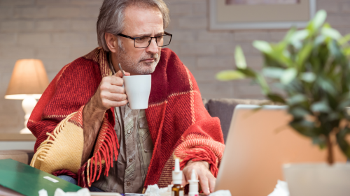 man huddled up in a blanket while looking at his laptop drinking tea