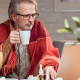 man huddled up in a blanket while looking at his laptop drinking tea