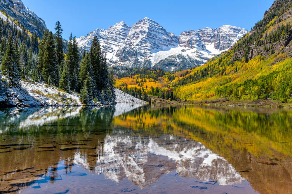 A wide-angle autumn midday view of snow coated Maroon Bells reflecting in crystal clear Maroon Lake